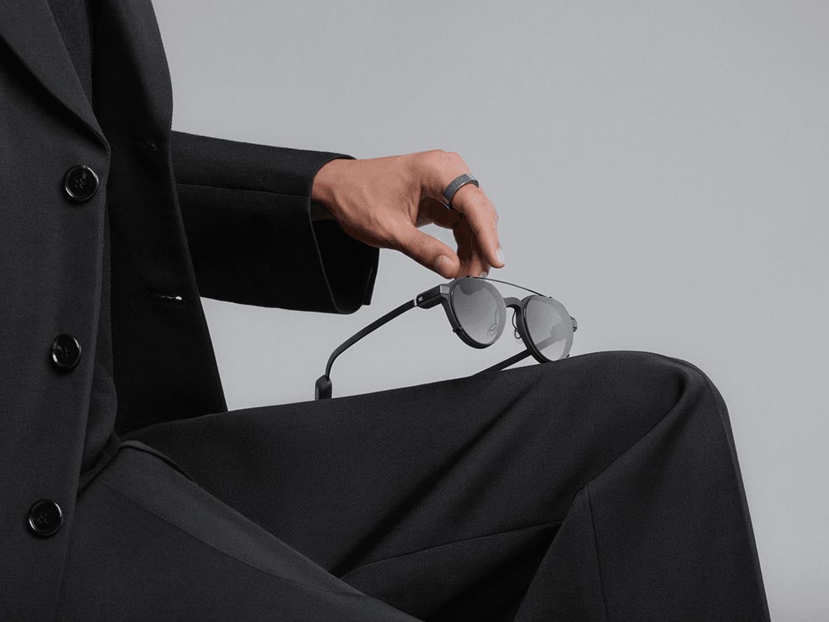 A hand holding minimal smart glasses while wearing a matching smart ring.