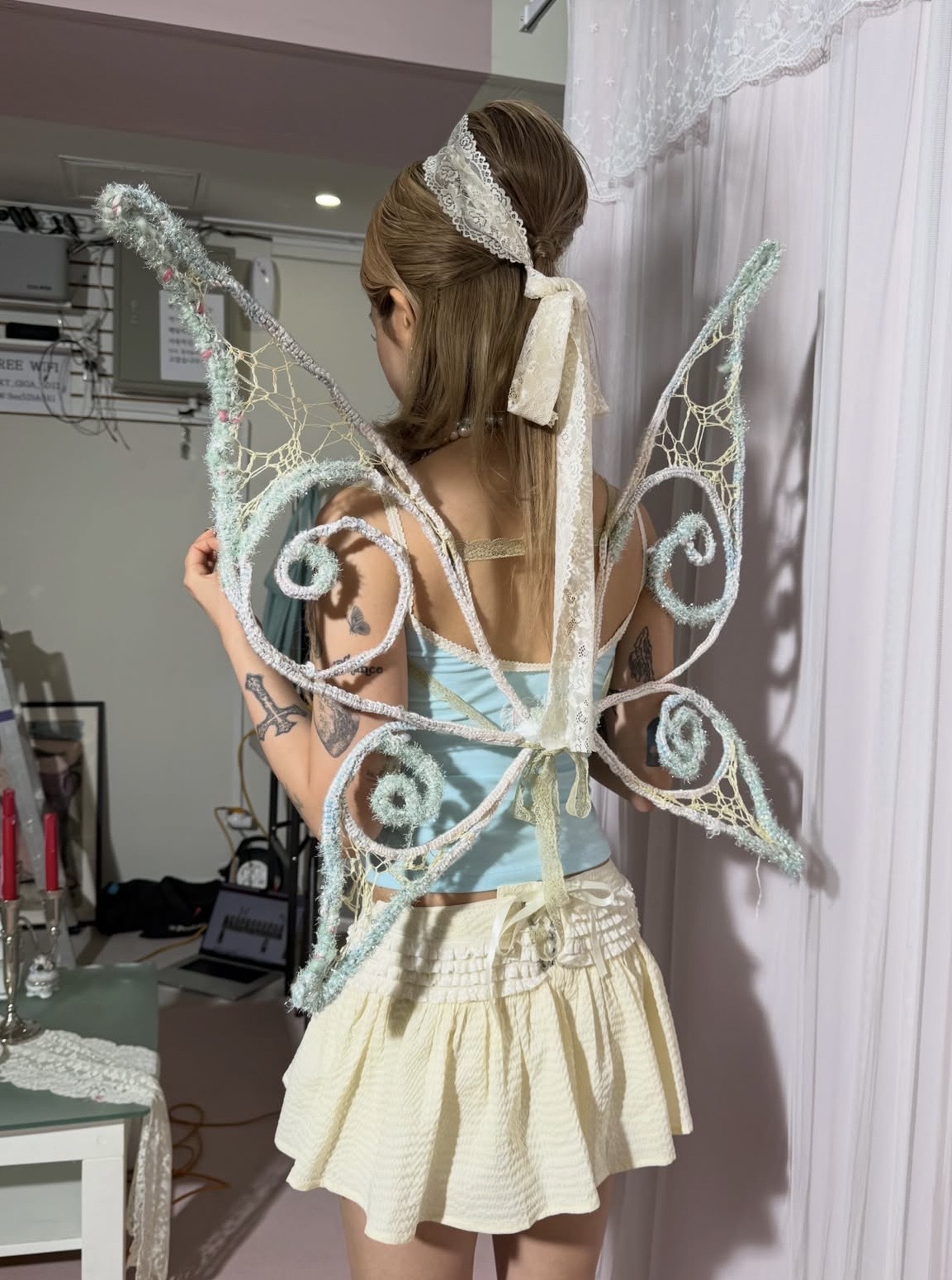 Back view of pale blue openwork fairy wings worn over a light blue top and cream skirt, with a lace hair bow.