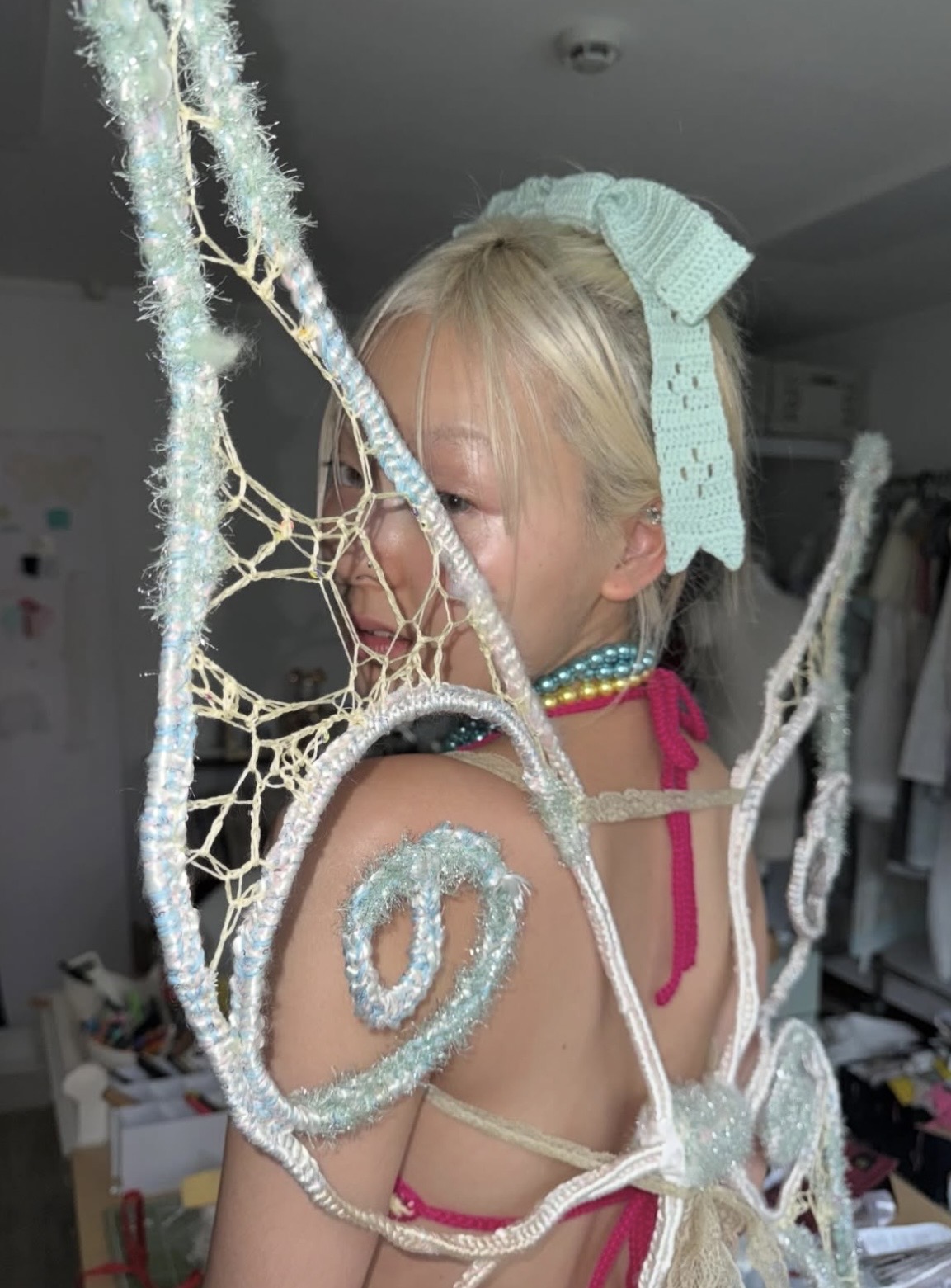 Close-up of a blonde model looking over her shoulder through curled pale blue wings, wearing a mint crochet bow.