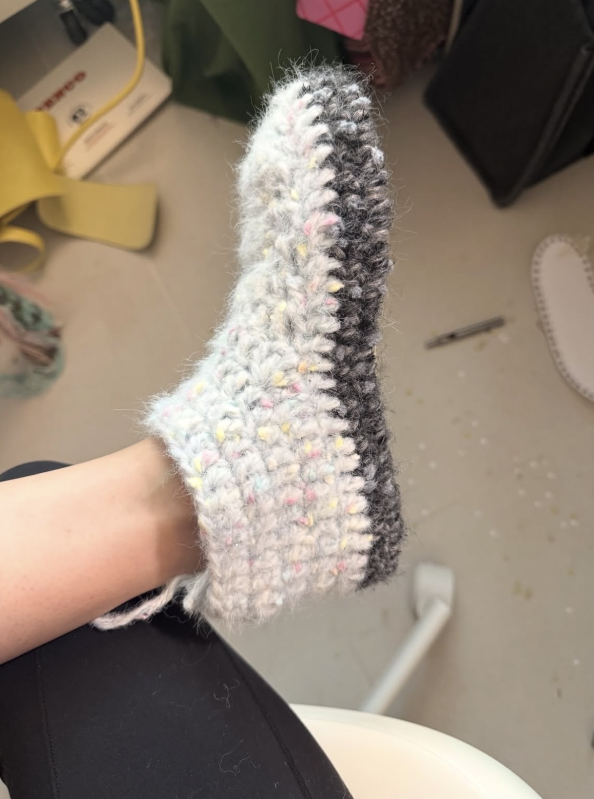 Side view of a chunky gray and pastel crochet slipper with a dark sole worn on one foot.