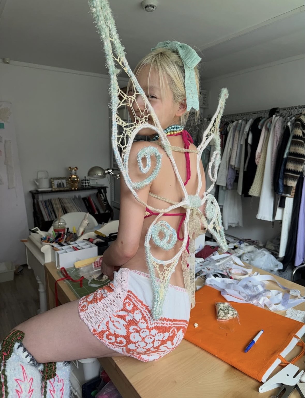 Seated model looking back through large curled wings, with a striped halter top and patterned white shorts.