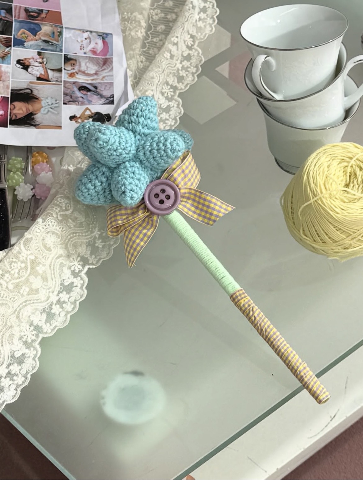 Crocheted blue star wand with a purple button and yellow gingham bow resting beside stacked cups and yarn.