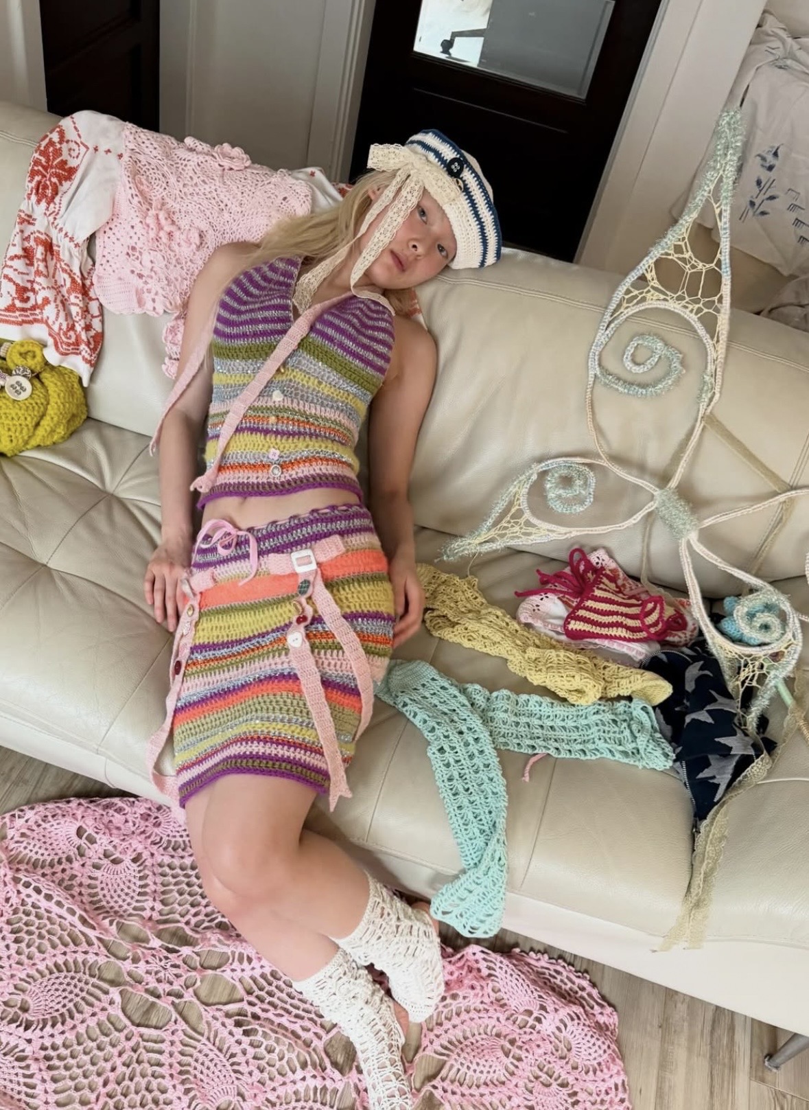Model reclining against a sofa in a striped crochet halter and skirt, with wings and pastel knitwear beside her.