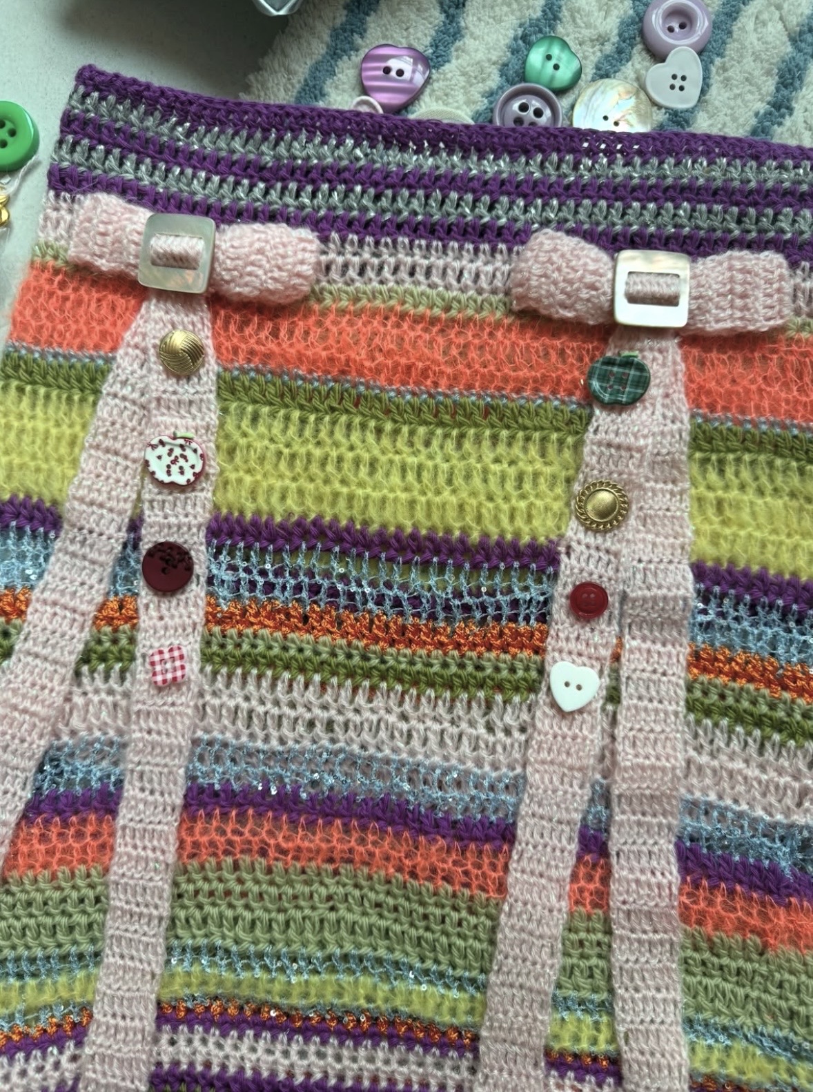 Detail of multicolored crochet stripes, pink straps and mismatched decorative buttons on a skirt.