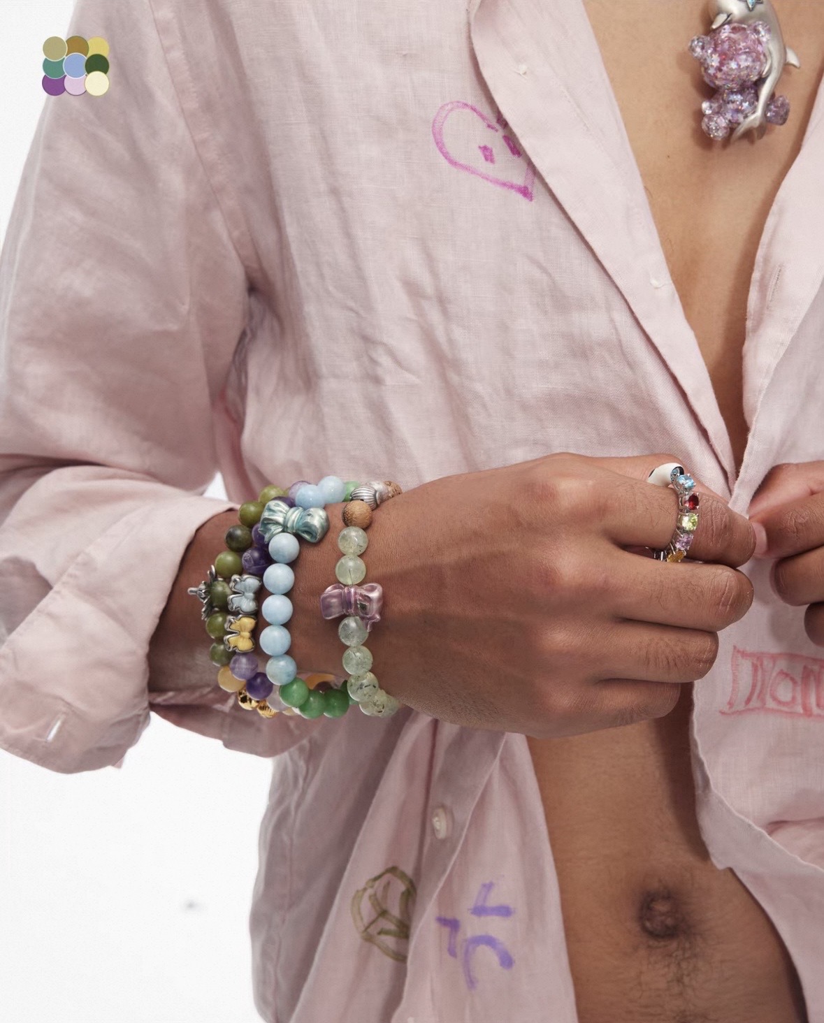 Several pastel and green bead bracelets stacked at the cuff of an open pale pink shirt, with decorative rings.