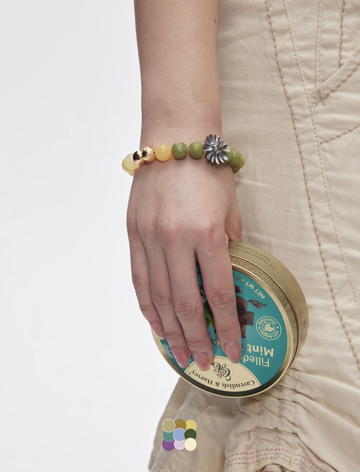 Yellow and green bead bracelet with a silver-colored flower charm on a hand holding a round tin.