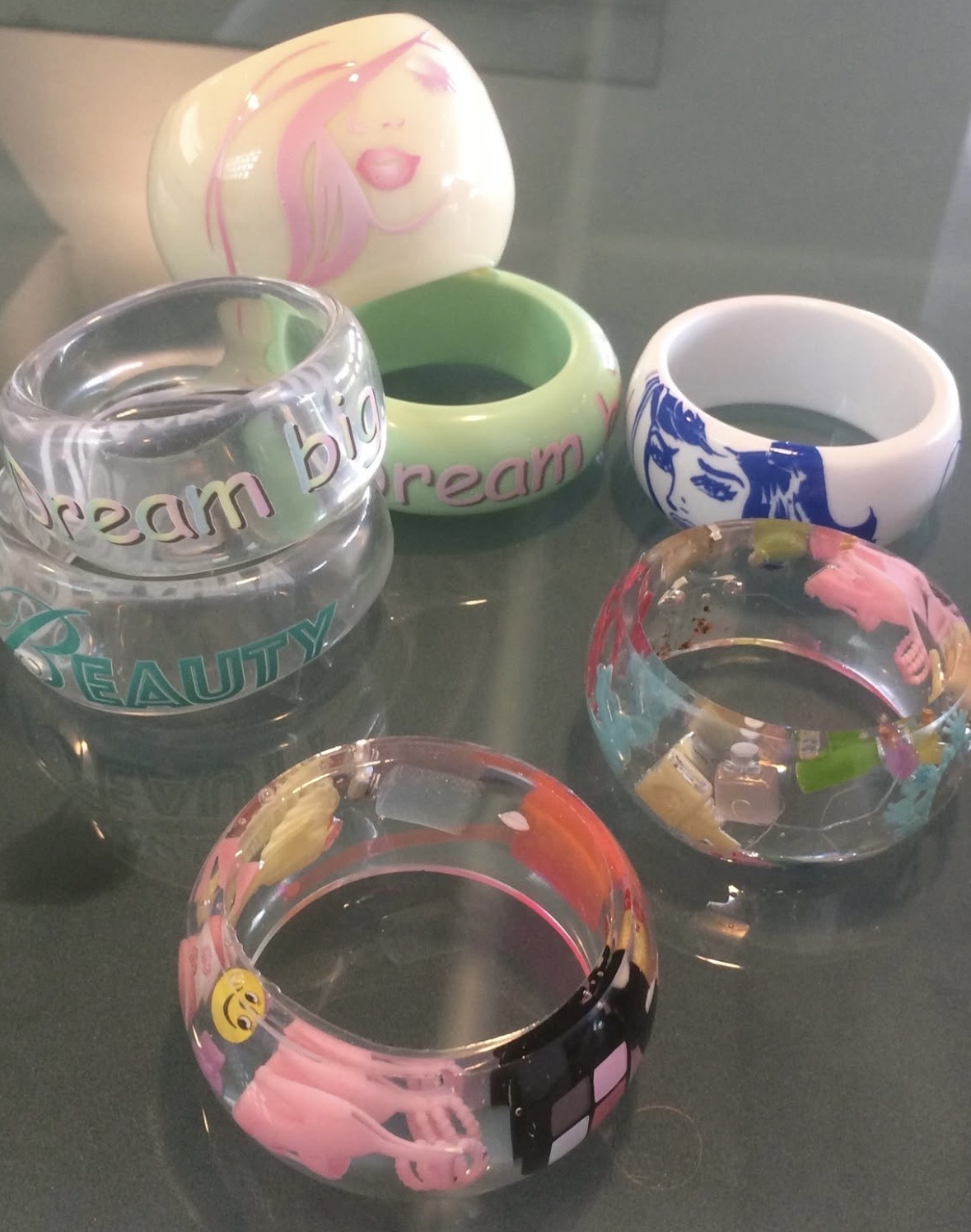 Assortment of wide pastel and clear bangles with lettering, illustrated faces and tiny embedded objects.