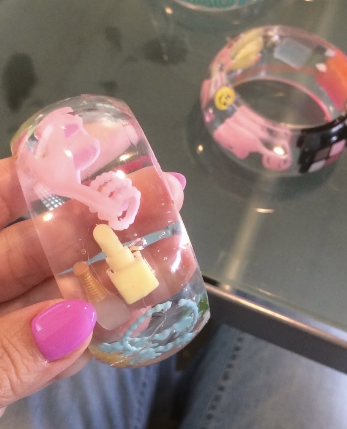 Hand holding a clear wide bangle filled with tiny pink, yellow and blue objects, with another cuff behind it.