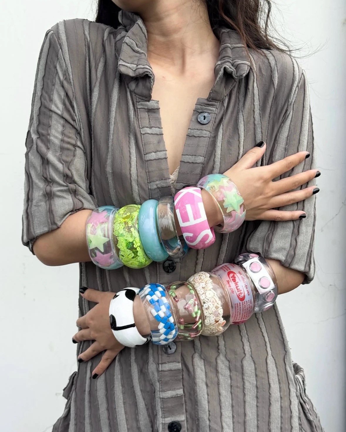 Person in a gray striped shirt wearing colorful chunky bangle stacks on both crossed arms.