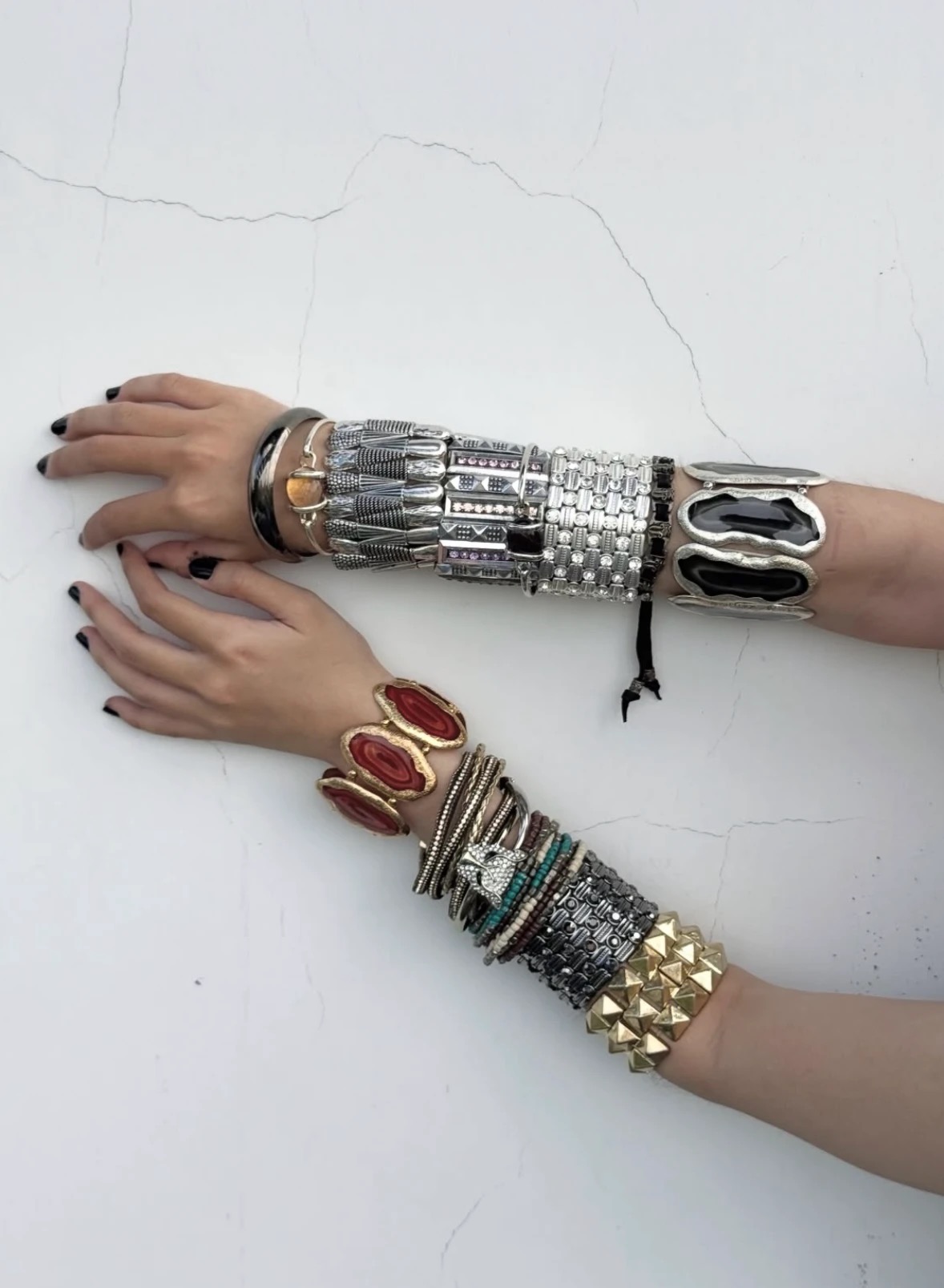 Two forearms against a white wall wearing stacked metallic bracelets, large red stones and a gold-colored studded cuff.