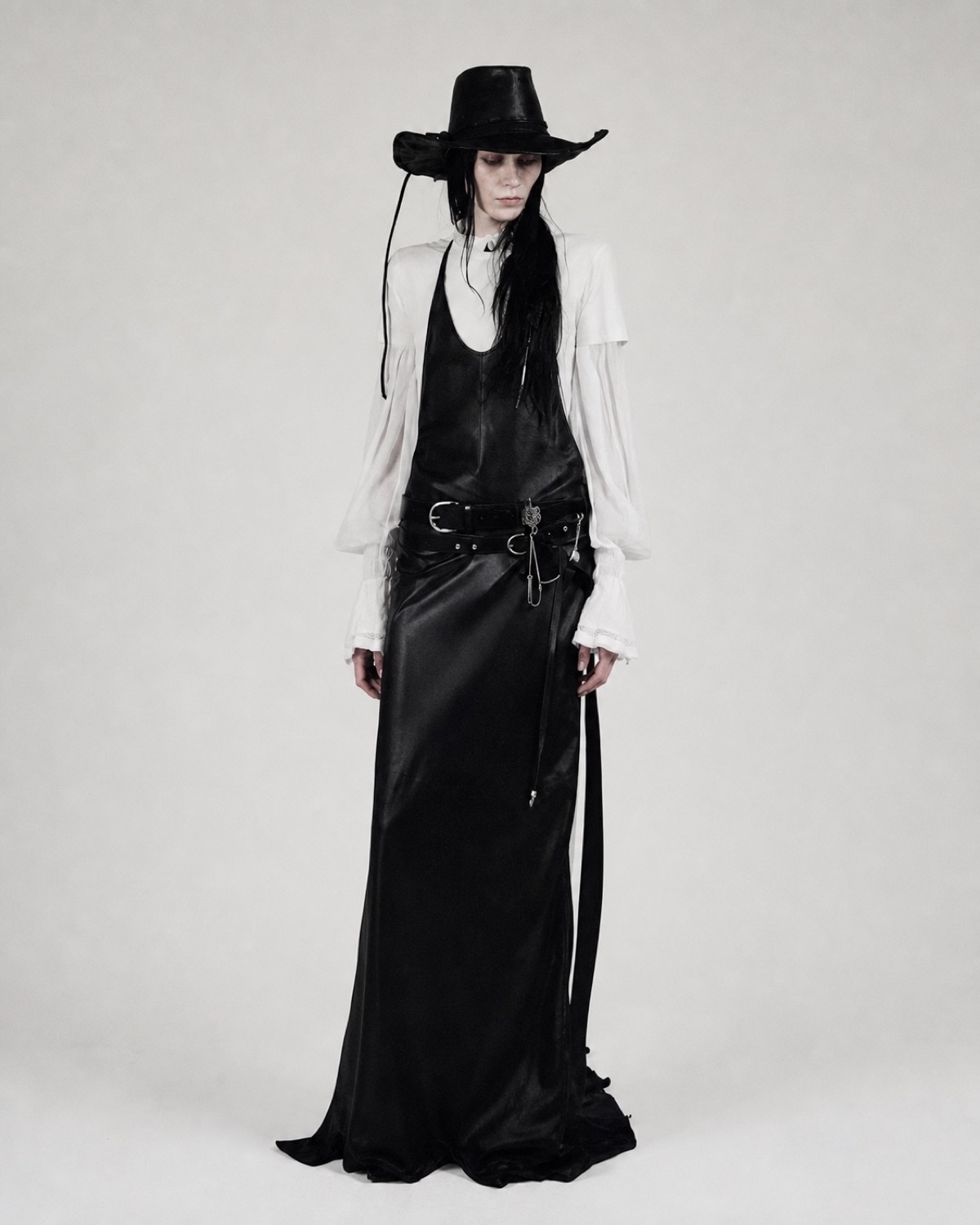 Sculptural black silhouette built through an oversized hat, leather layers and a floor-length skirt