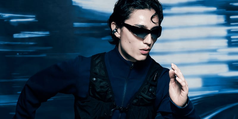 Model wearing open-ear audio with dark eyewear and a technical jacket in a blue motion-lit scene.