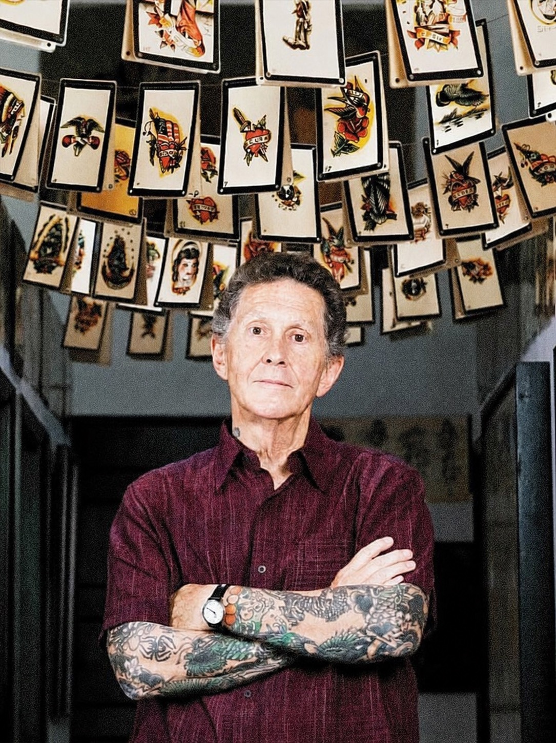 A portrait of Don Ed Hardy standing beneath tattoo flash artwork.