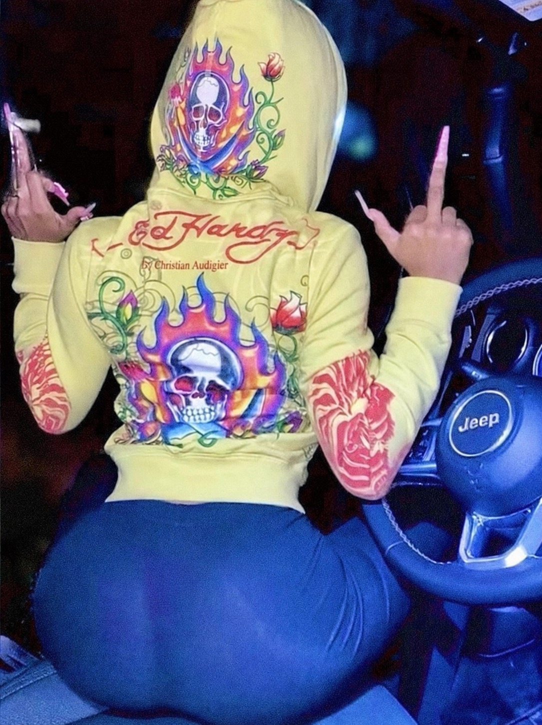 A yellow Ed Hardy hoodie with skull, flame, and rose graphics worn in a car.