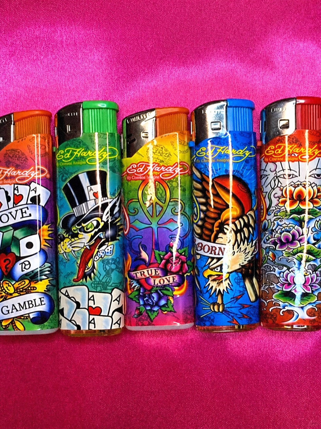 A row of colourful Ed Hardy lighters with tattoo-style graphics on a hot pink background.