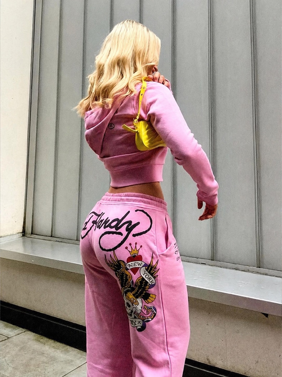 A pink Ed Hardy tracksuit styled from the back with a yellow shoulder bag.