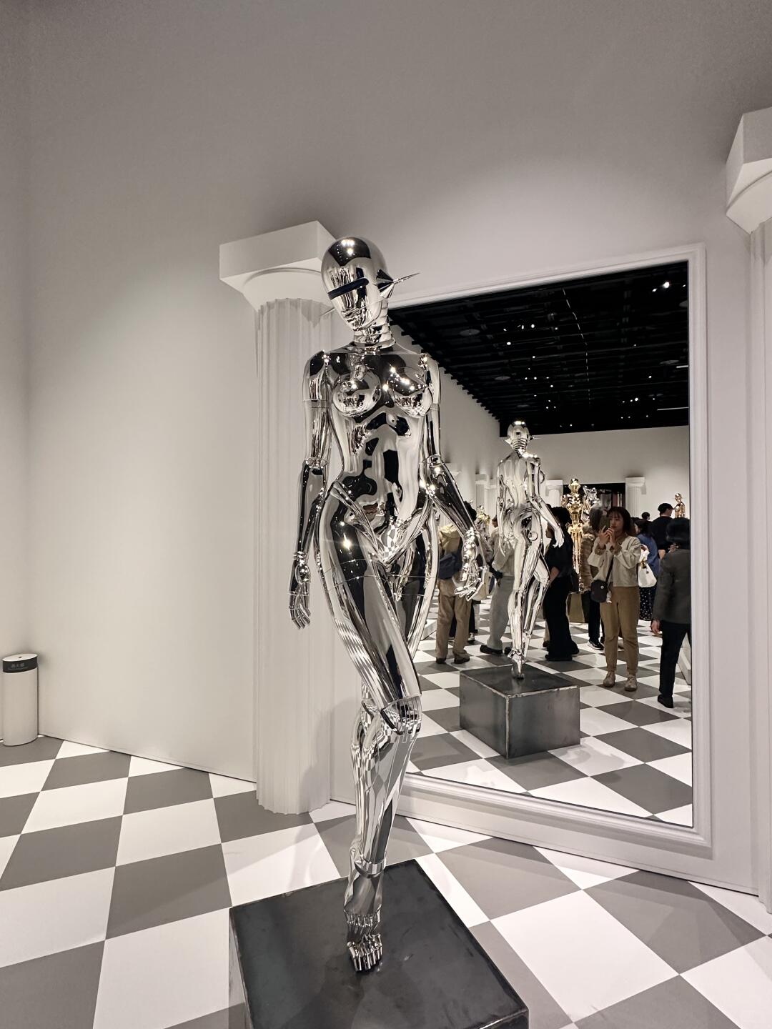 Life-size silver chrome gynoid standing in a gallery with checkerboard floor