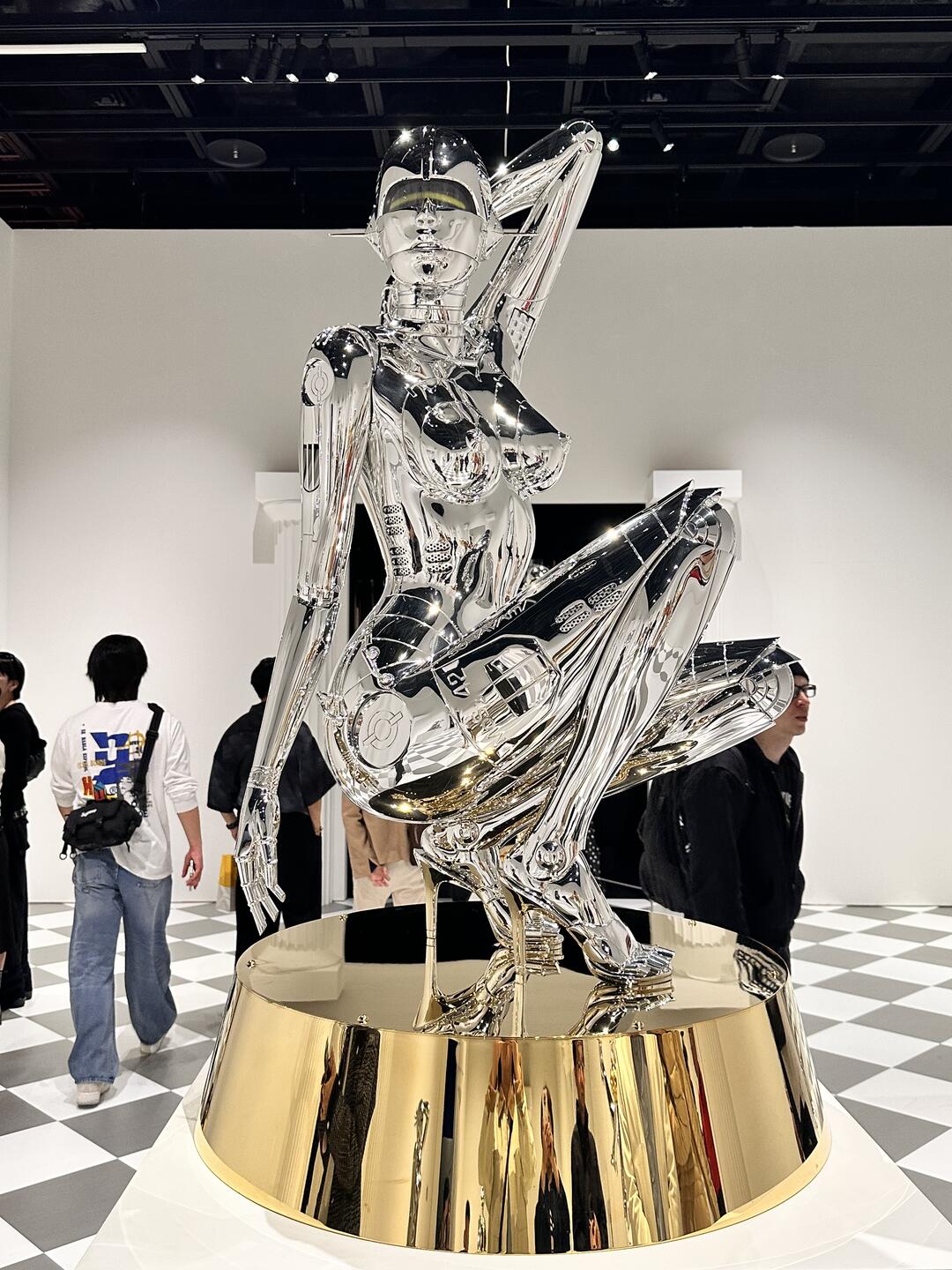 Large chrome gynoid sculpture posed on gold plinth in exhibition setting