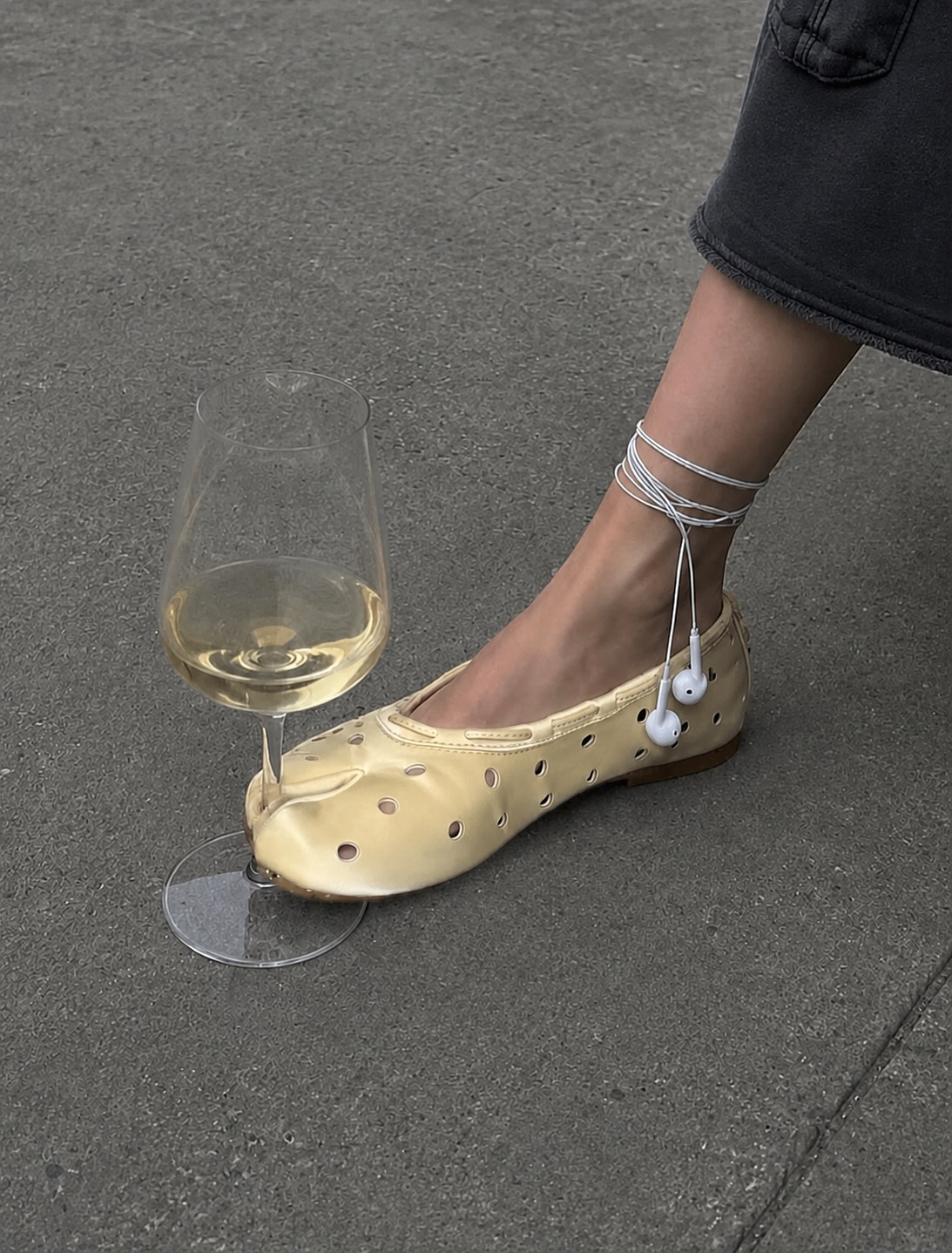 A cream mule flat styled with a wired earbud and a wine glass on grey pavement.