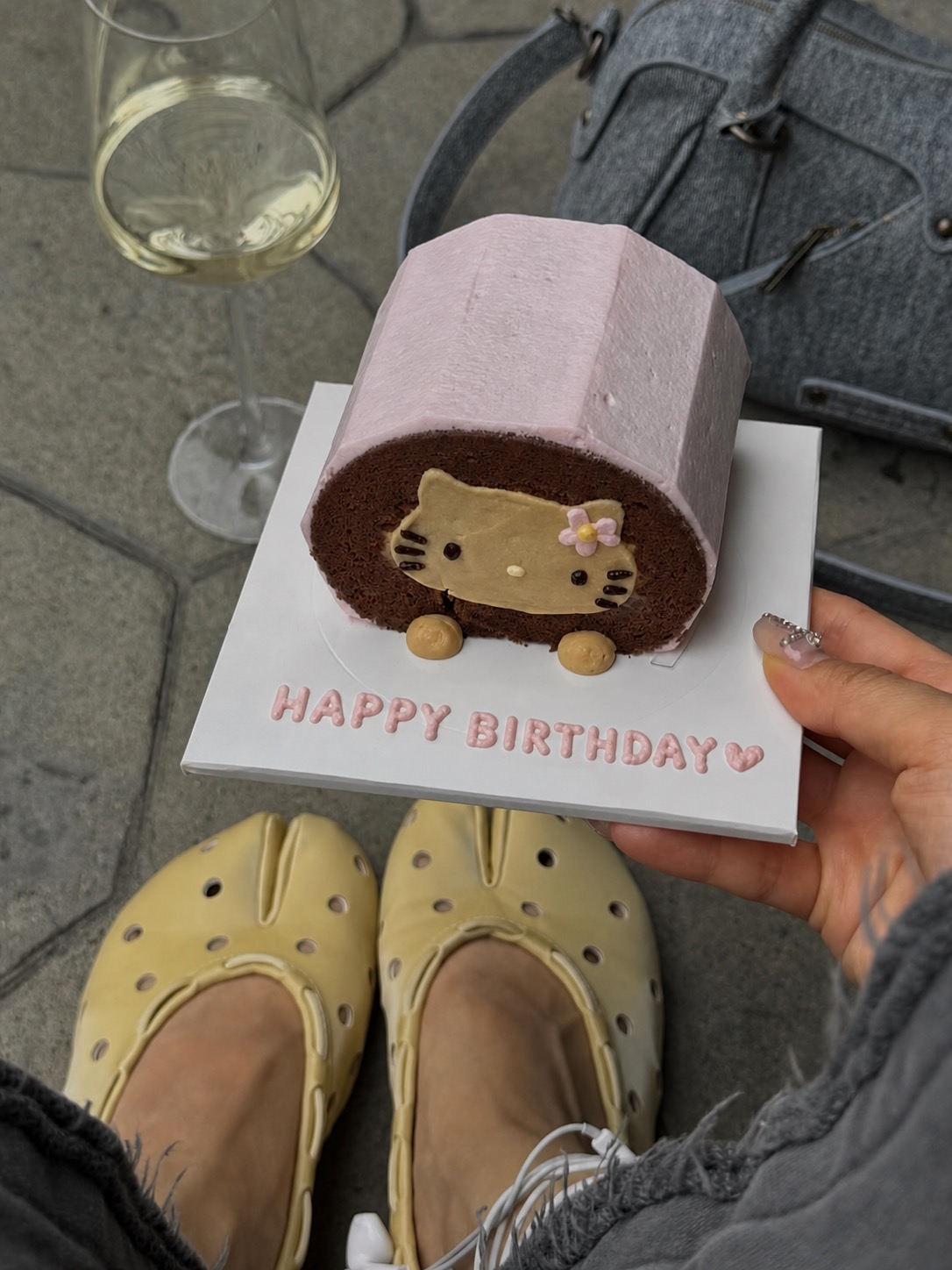 Cream mule flats beside a birthday cake, a wine glass, and a grey handbag.