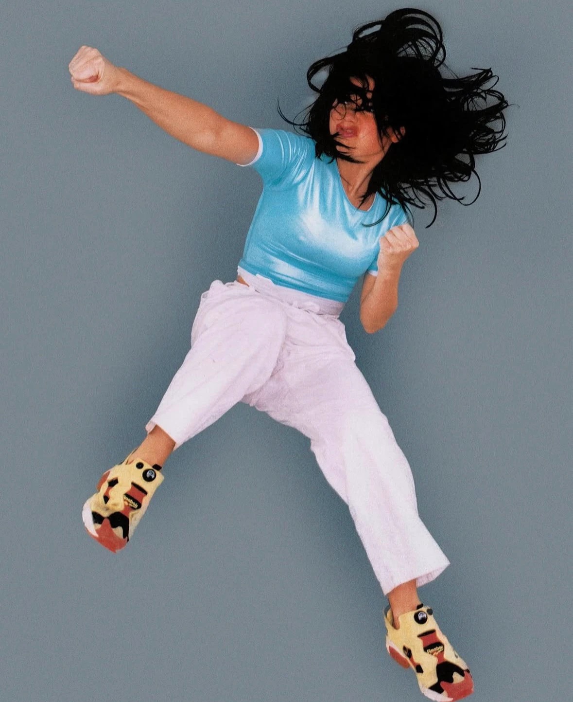 Airborne model wearing a blue T-shirt, pale-pink cropped trousers and yellow-and-red cutout sneakers against a gray backdrop.