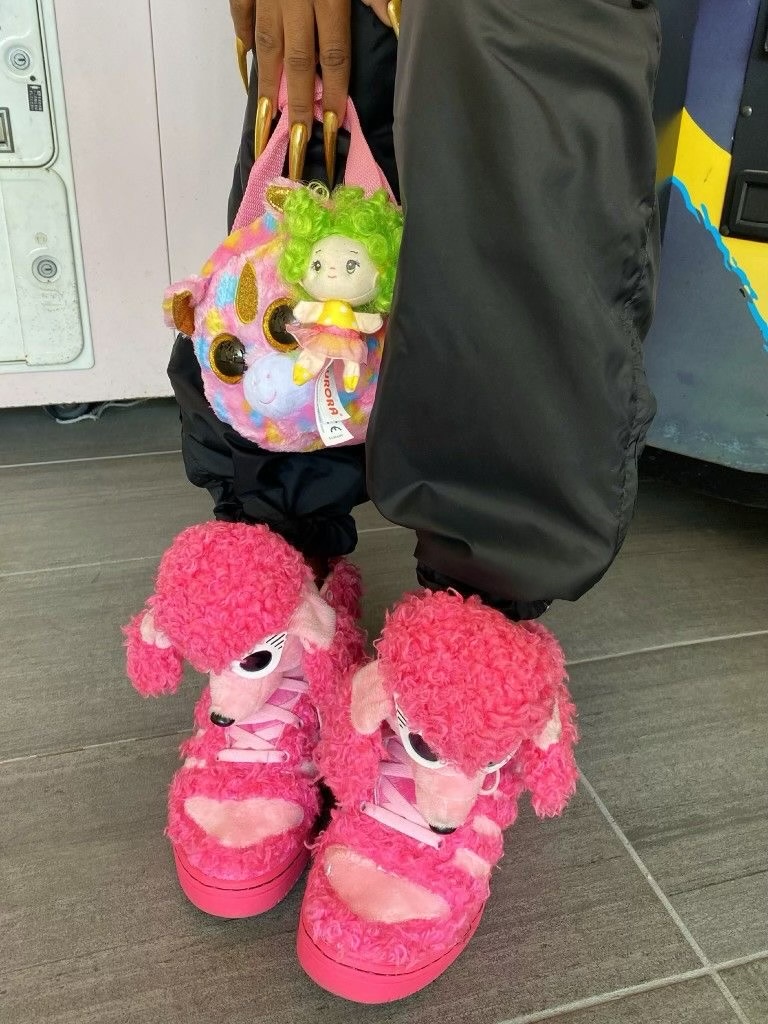 Bright pink poodle-head high-tops beneath loose black trousers, beside a colorful plush bag with a dangling doll.