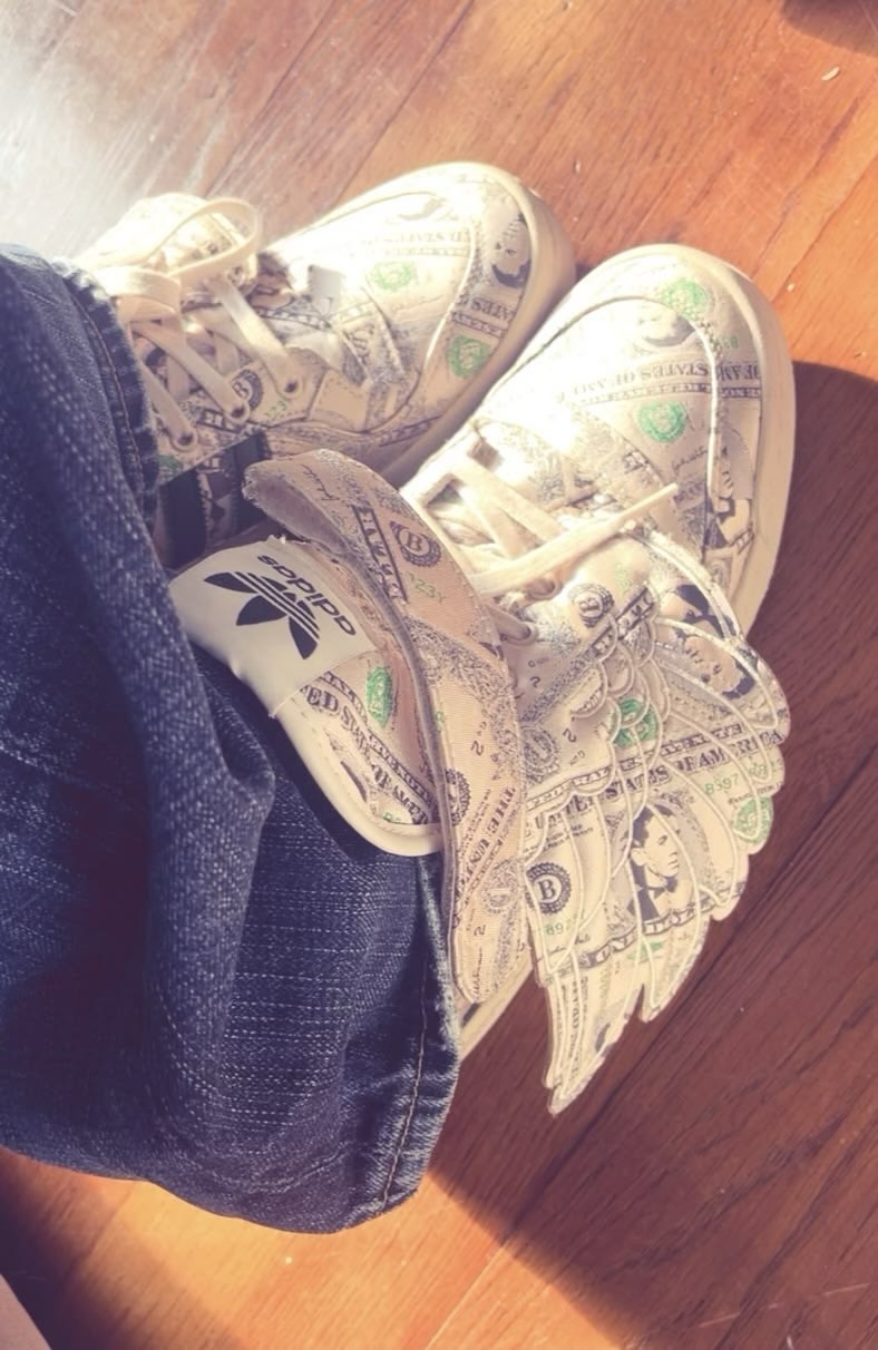 Close-up of banknote-print winged sneakers worn with dark jeans on a wooden floor.