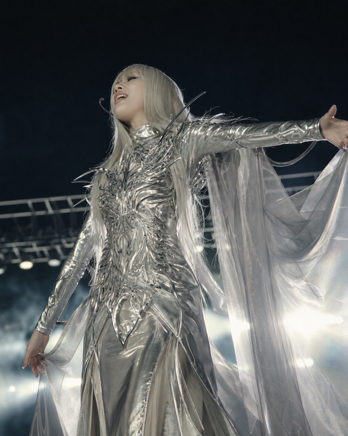 A fictional blonde stage performer in a silver chrome angel outfit with a translucent cape under night concert lights.