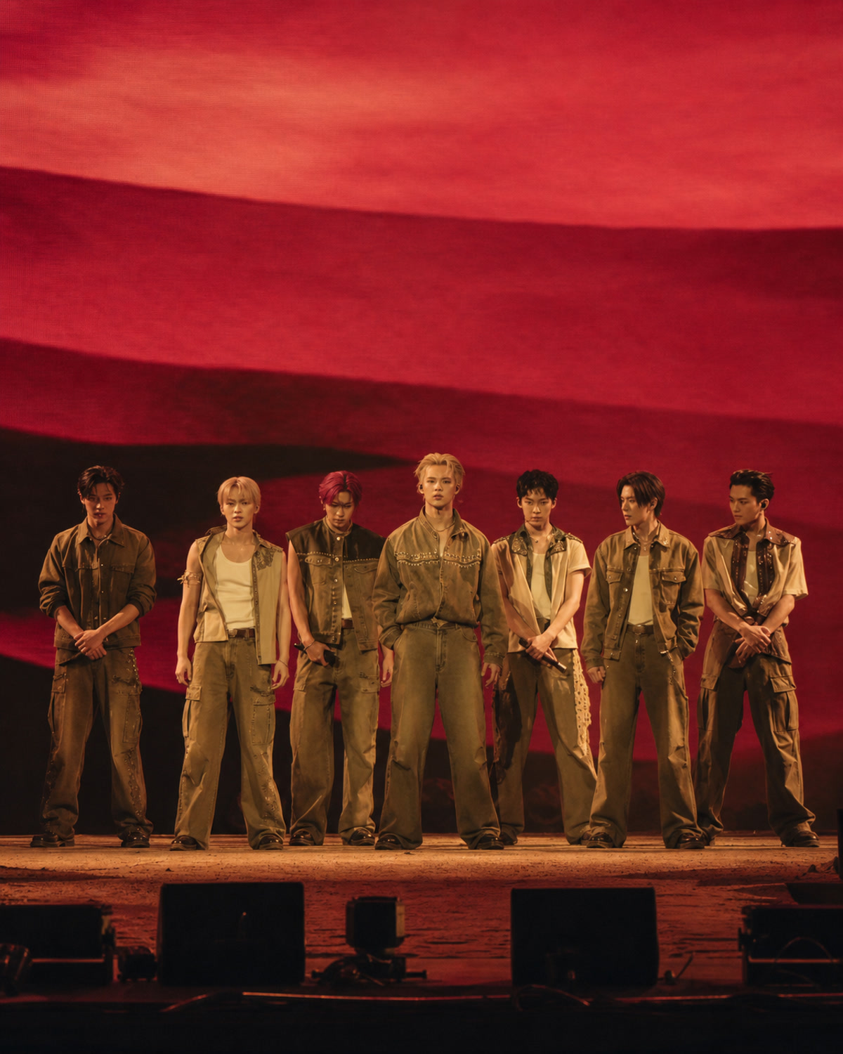 Seven fictional male performers in coordinated tan denim stage outfits standing before a red desert concert backdrop.