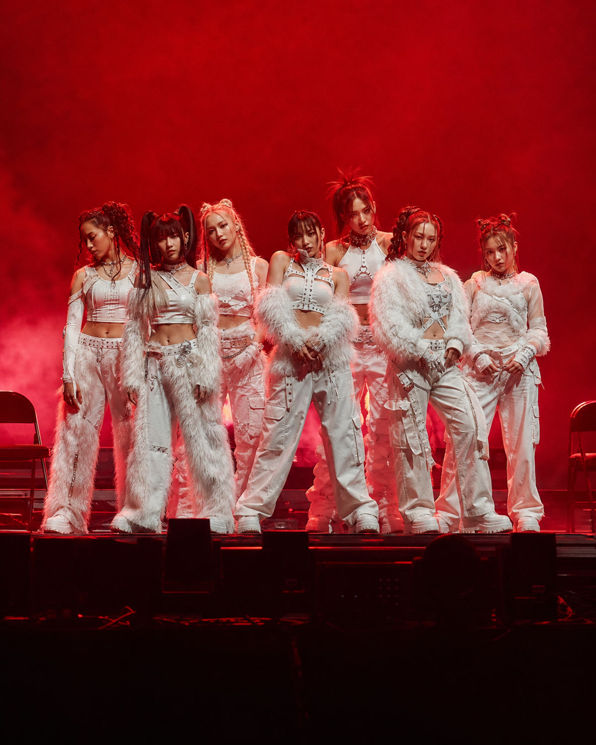 Seven fictional female performers in white futuristic stagewear holding a tight red-lit concert formation.