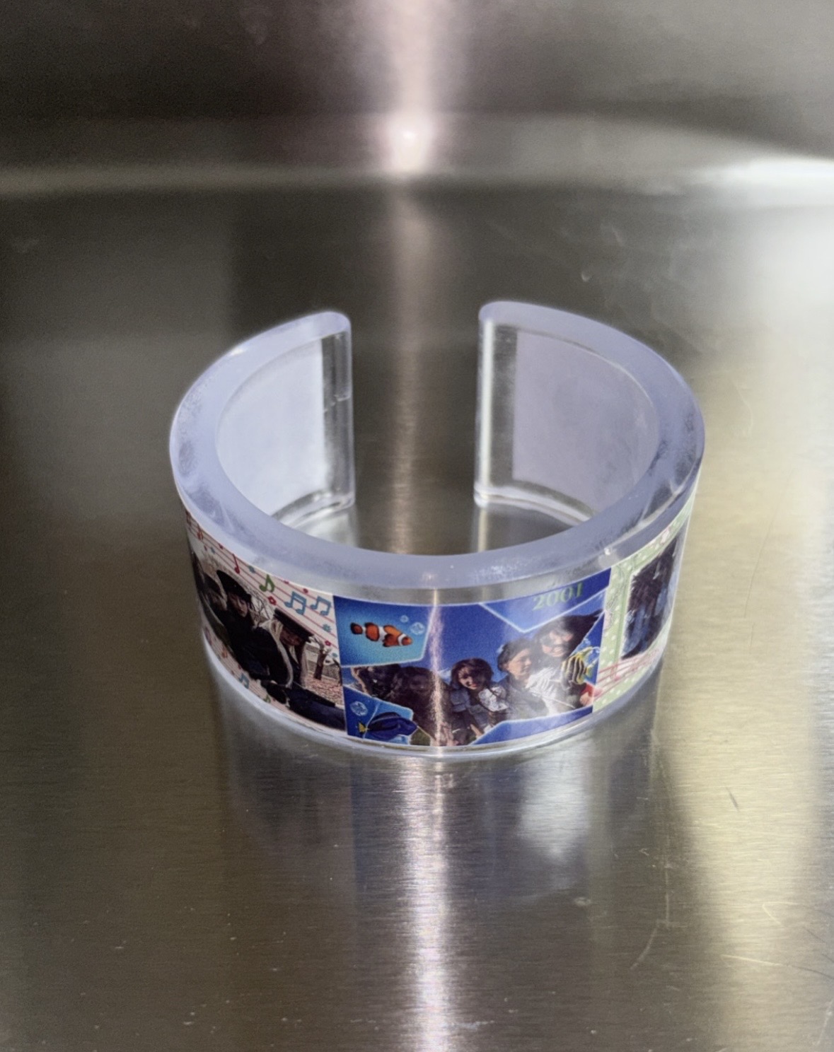 Clear open cuff with a blue-toned photo collage resting on a reflective metal surface.