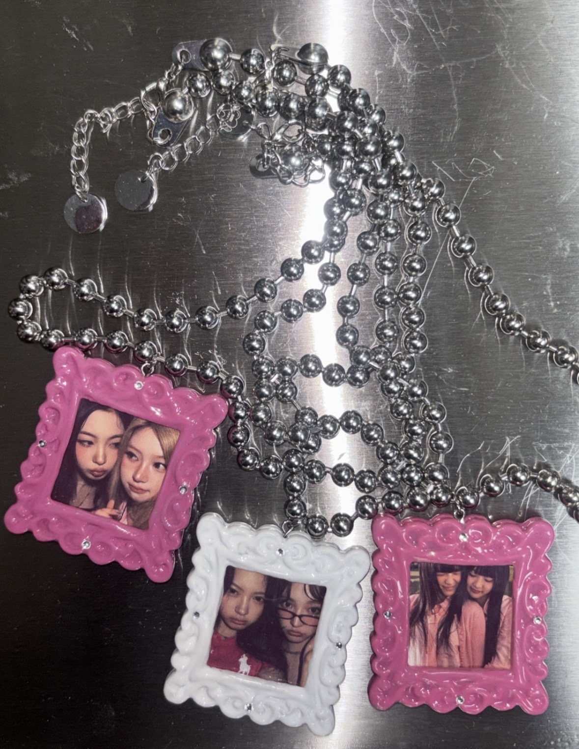 Pink and white miniature photo-frame pendants on silver-colored chains laid across a metal surface.