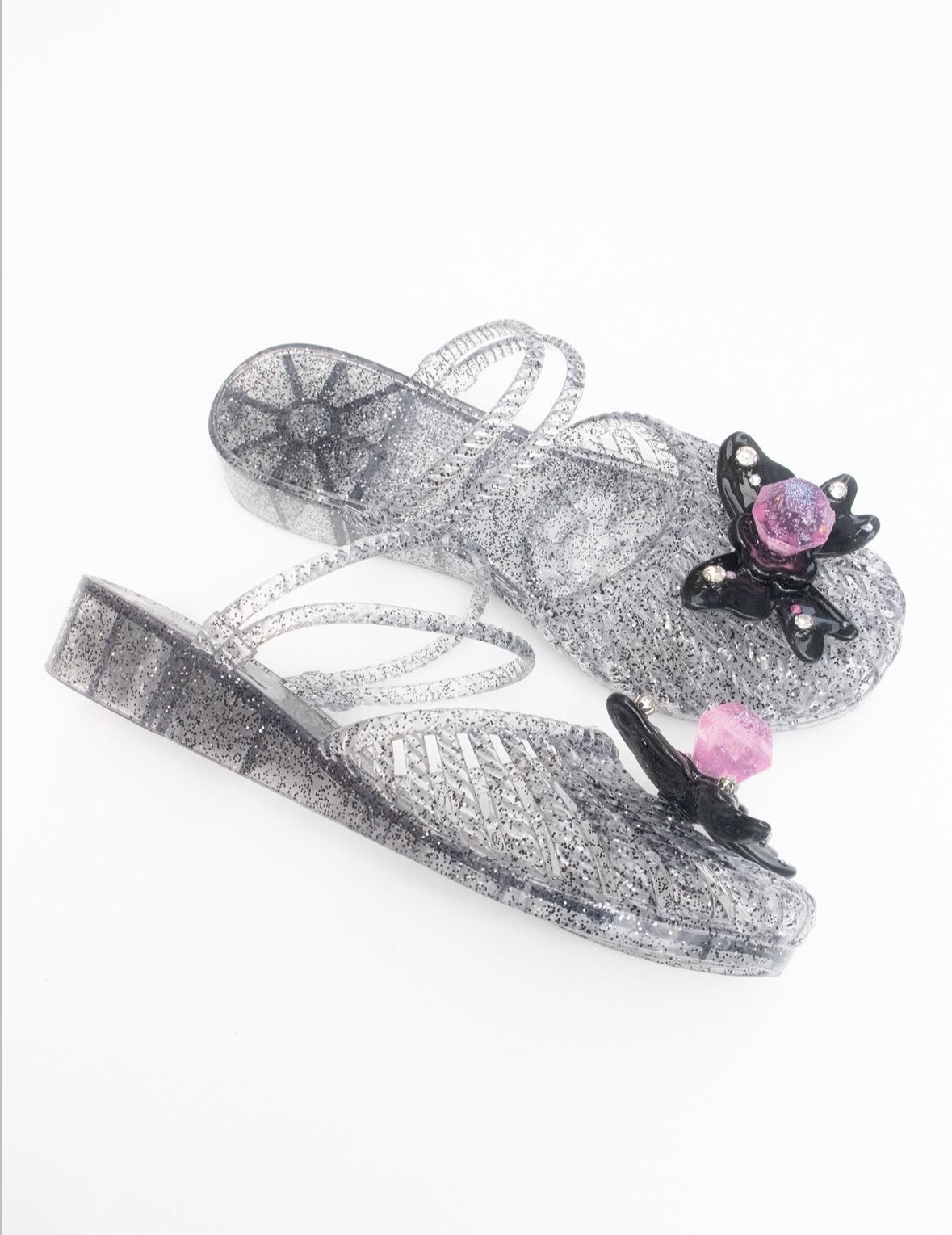 Pair of clear glitter heeled sandals decorated with black wing-like shapes and pink faceted ornaments.