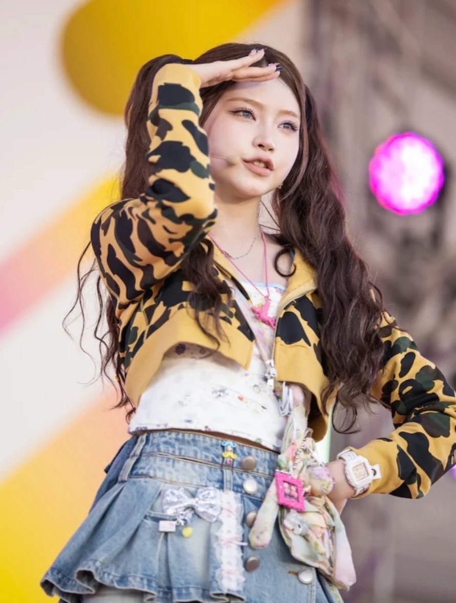 Performer in a leopard-print cardigan and denim skirt wearing a pink photo-frame pendant among layered charms.