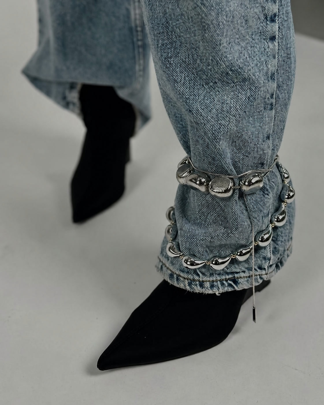 Heavy silver chain anklet gathered around light denim and a black pointed shoe