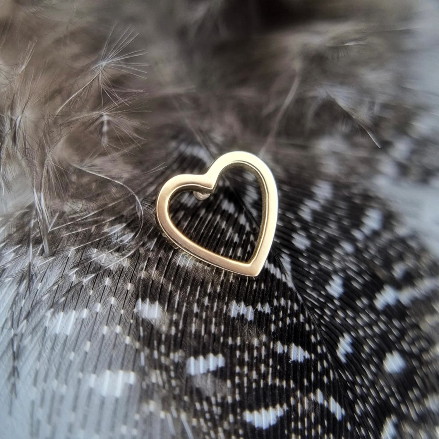 AURIS gold heart piercing piece photographed against dark feathers