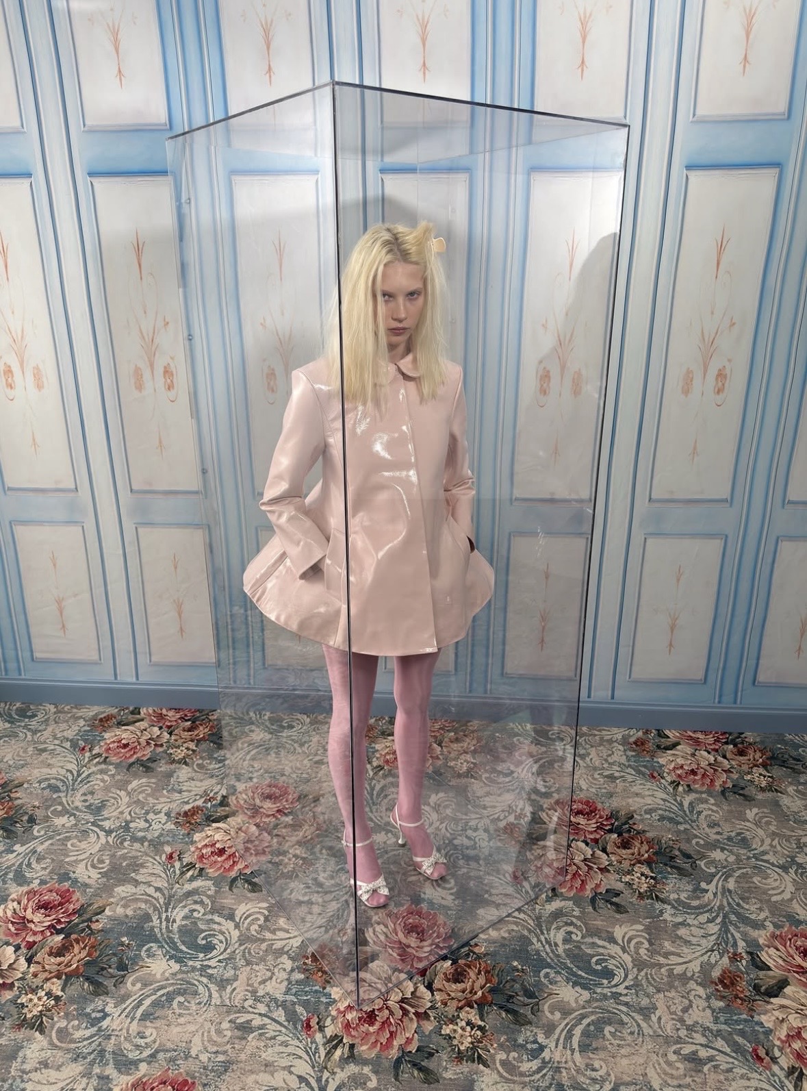 Model in a glossy pale-pink sculptural jacket and pink tights, standing inside a transparent display case in a blue-paneled room.