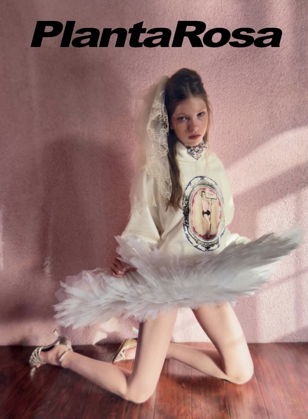 Planta Rosa campaign image of a kneeling model in a cream graphic sweatshirt, lace head covering and flared pale tulle skirt.