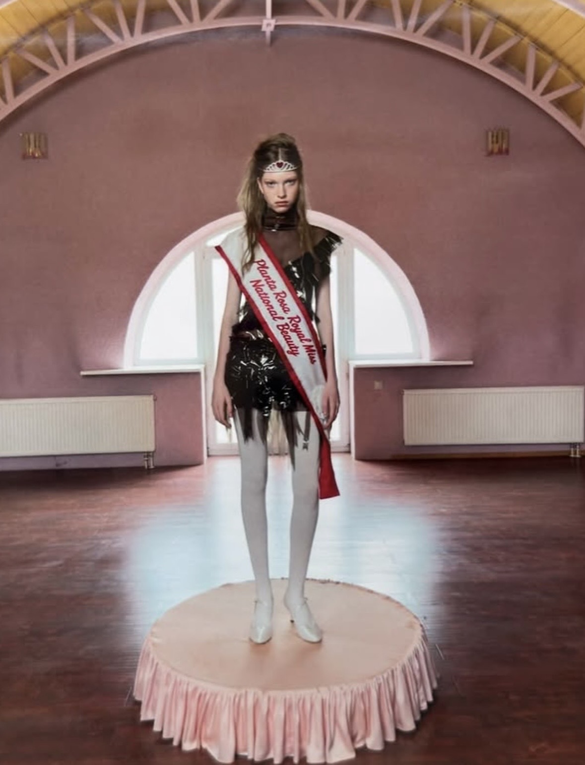 Model wearing a tiara, a white-and-red pageant sash, a dark sculptural mini dress and white tights on a round pink platform.