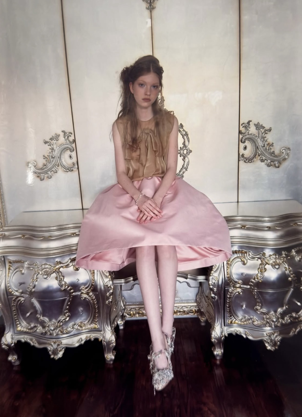 Seated model in a tan ruffled sleeveless top, a full pale-pink skirt and embellished shoes against an ornate cabinet.