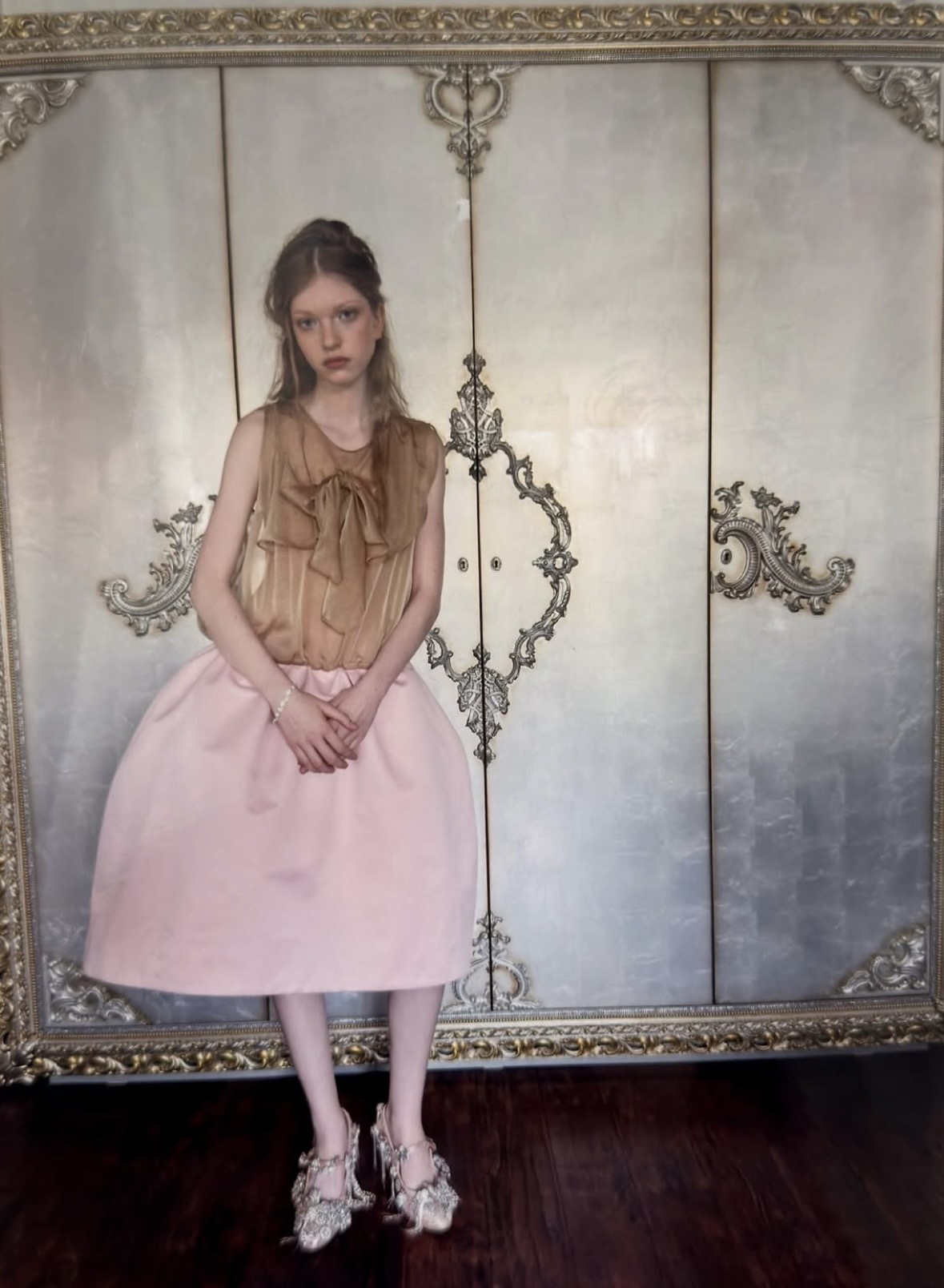 Standing view of a model in a tan tie-neck top and structured pink skirt, with silver-colored embellished shoes.