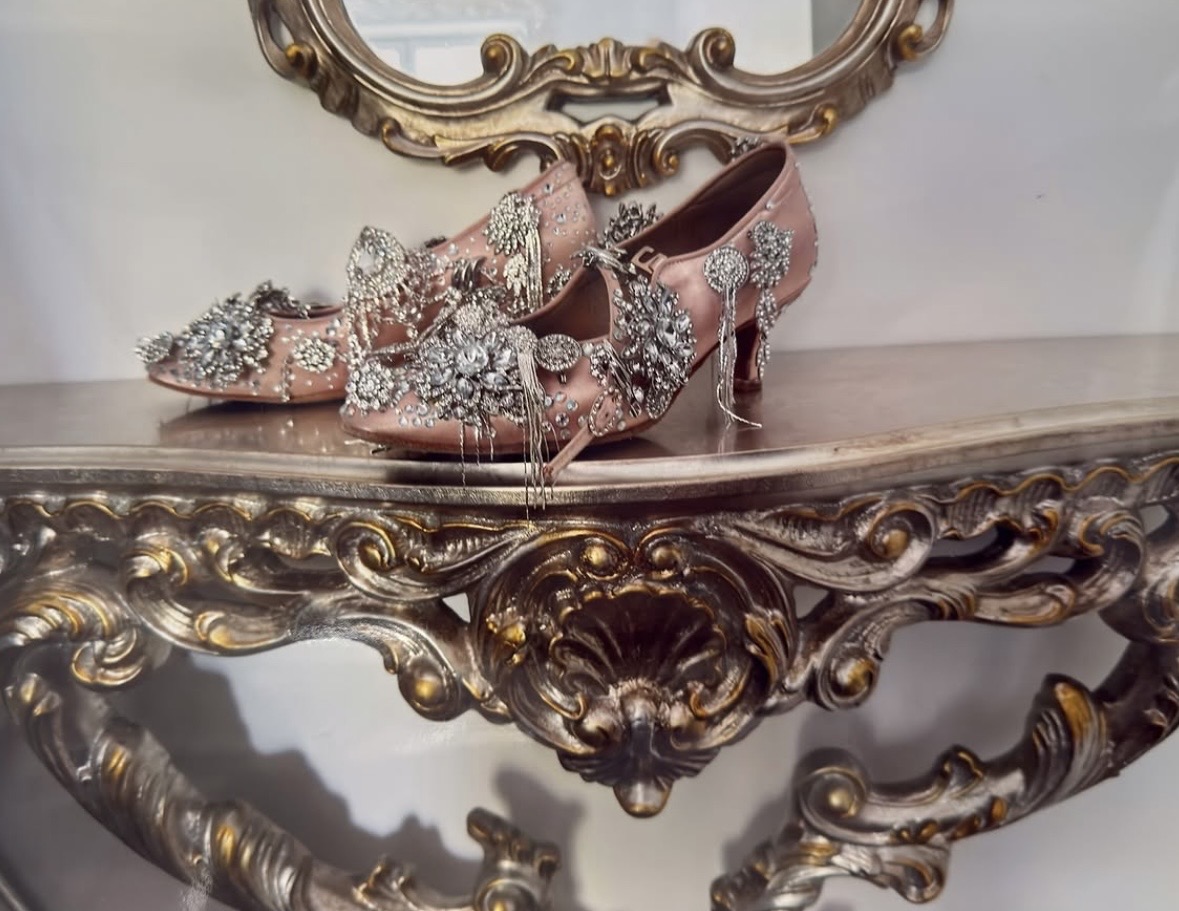 Pink heeled shoes covered in sparkling ornaments and dangling chains, displayed on an ornate gold-colored console.