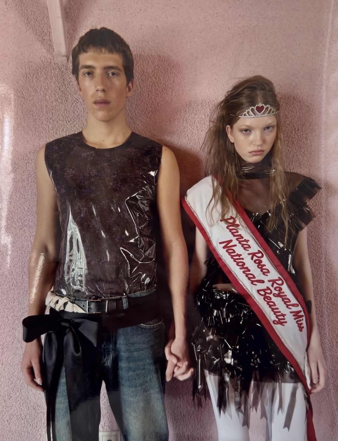 Two models holding hands in dark glossy outfits; one wears jeans with a large black bow, and the other wears a tiara and pageant sash.