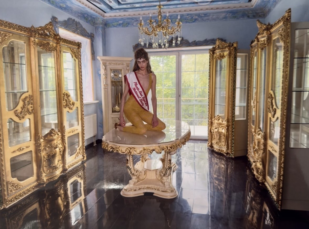 Model in a mustard-yellow outfit and pageant sash kneeling on a table between gilded display cabinets beneath a chandelier.