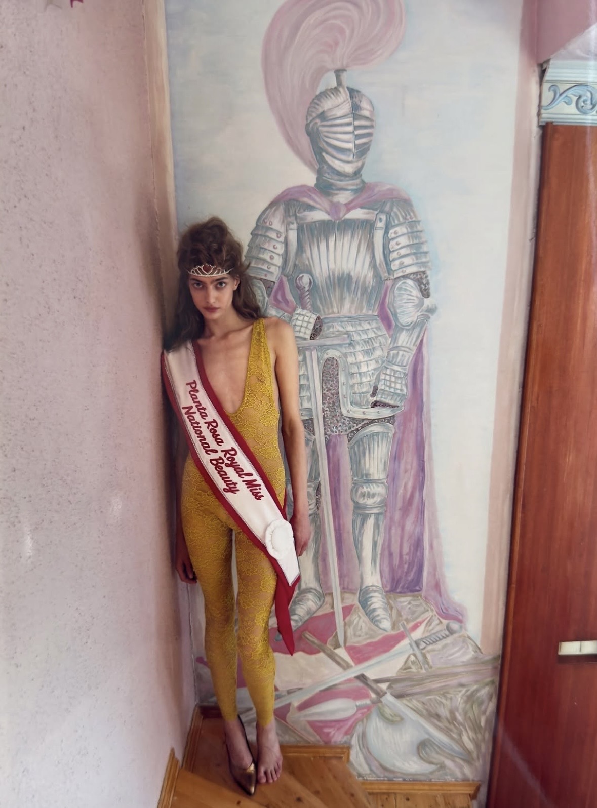 Barefoot model in a mustard-yellow plunging one-piece, tiara and pageant sash beside a mural of an armored figure.