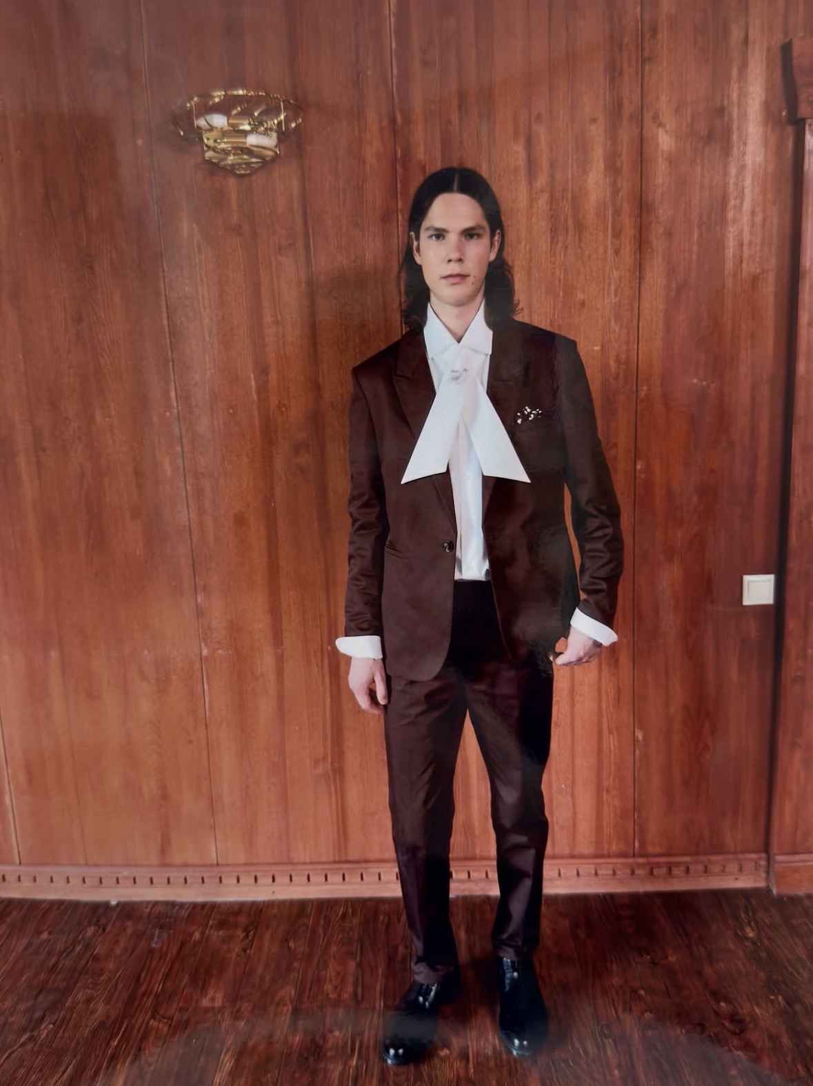 Model in a dark brown suit with a long white neck bow, white shirt cuffs and black shoes against a wood-paneled wall.