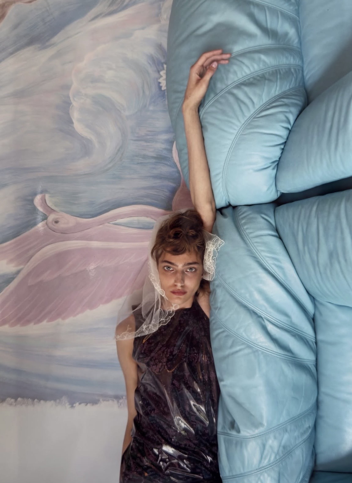 Model wearing a dark glossy sleeveless top and lace head covering, reclining beside pale-blue padded seating and a painted wall.