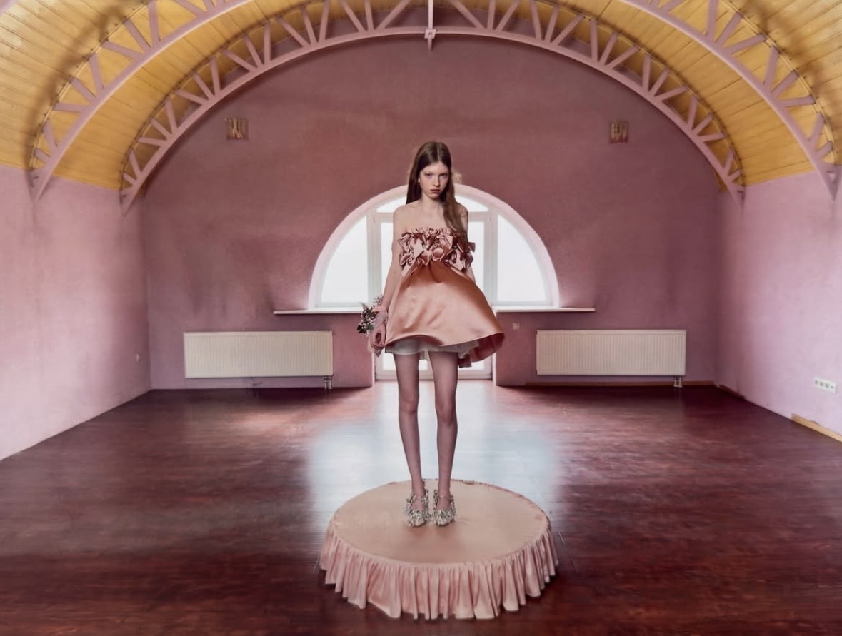 Model in a pale-pink mini dress with large floral details and a flared skirt, standing on a circular platform in a pink hall.