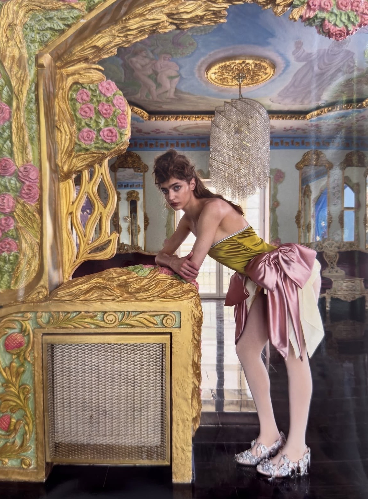 Model in a chartreuse strapless outfit with an oversized pink bow, leaning against a painted decorative panel beneath a chandelier.