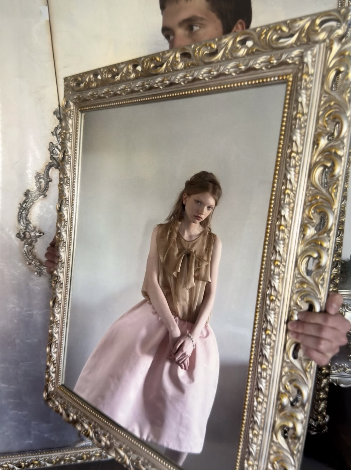 A large ornate mirror held upright reflects a model wearing a tan ruffled top and pale-pink skirt.
